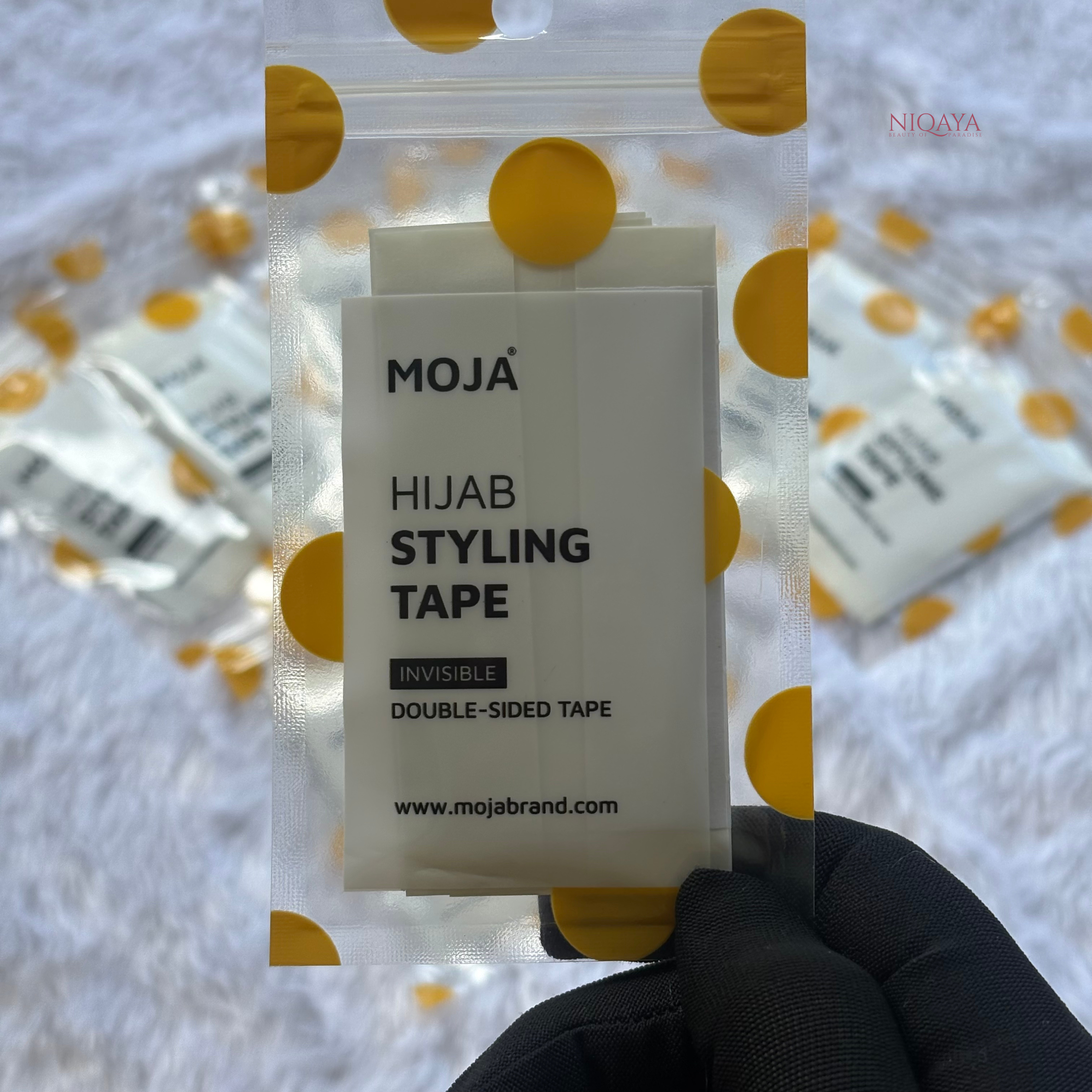 MOJA Hijab Styling Tape packaging held in front of a blurred background with yellow circles.