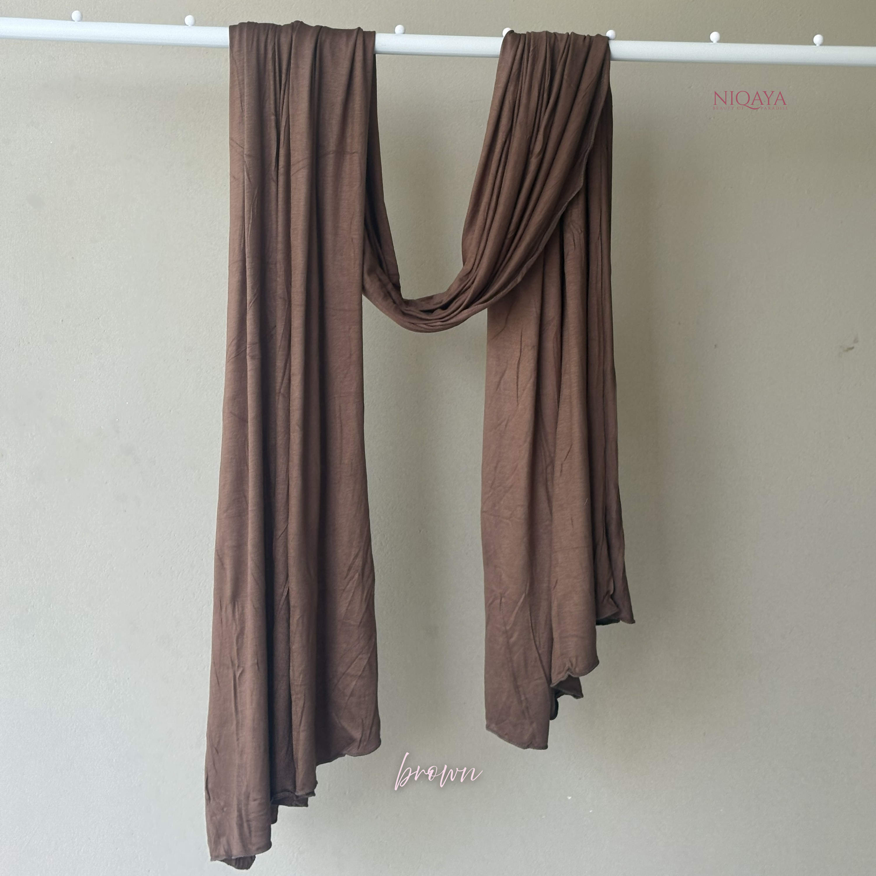 Brown scarf draped over a white rod against a beige wall with 'NIQAYA' branding.