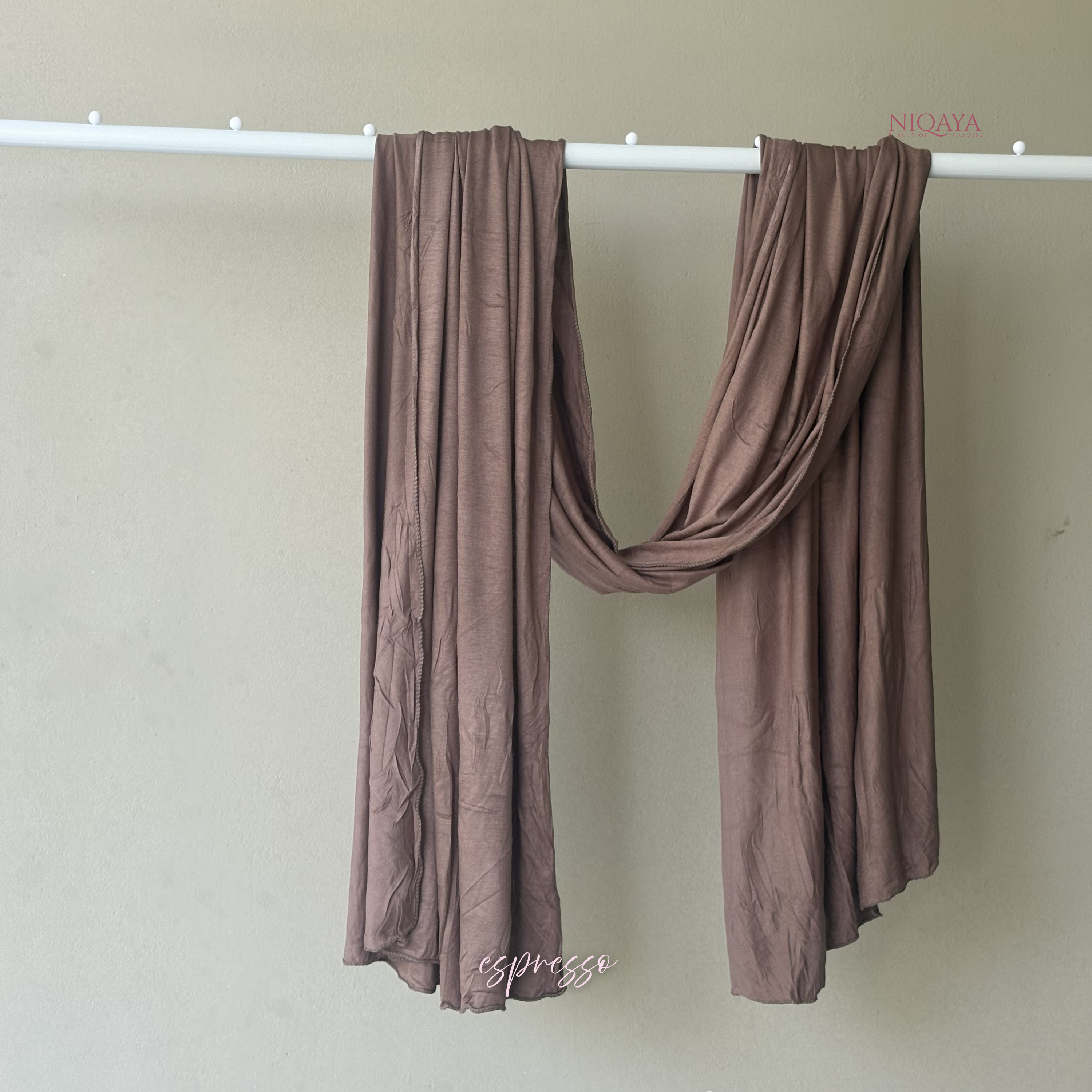 Brown scarf draped over a white rod against a beige wall with 'Niqaya' branding.